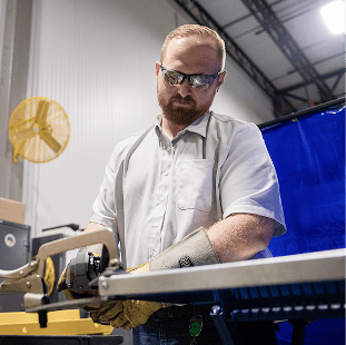 A person wearing safety glasses and gloves uses a saw to cut metal in an industrial workshop setting.