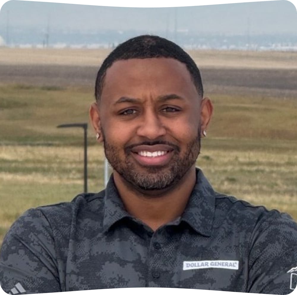 A man with short hair and a beard smiles outdoors. He is wearing a gray patterned shirt with a “Dollar General” logo. The background shows an open field.