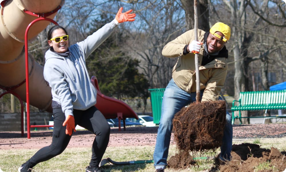 Two people outdoors, smiling while digging and moving soil. One holds a shovel, the other poses playfully. Both wear gloves. Playground equipment and benches are visible in the background on a sunny day.