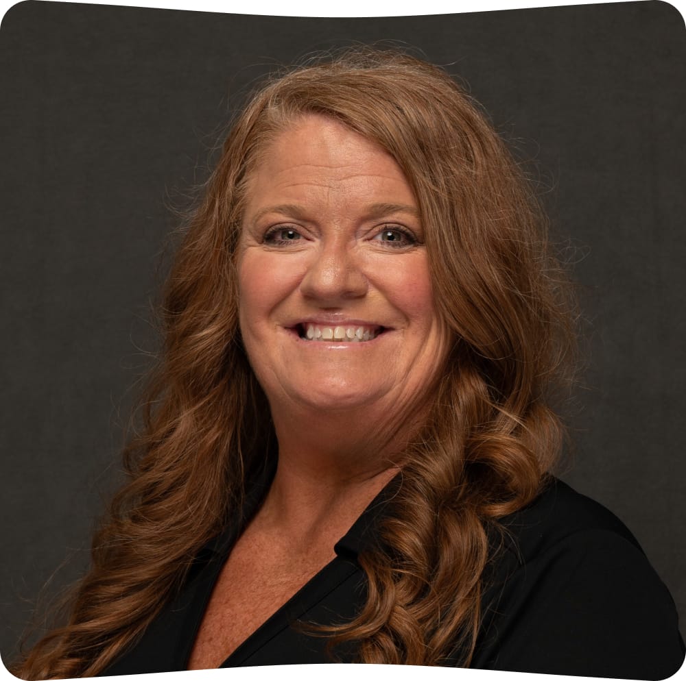 Woman with long wavy light brown hair smiles at the camera. She is wearing a black top and is posed in front of a dark gray background. The image has rounded corners.