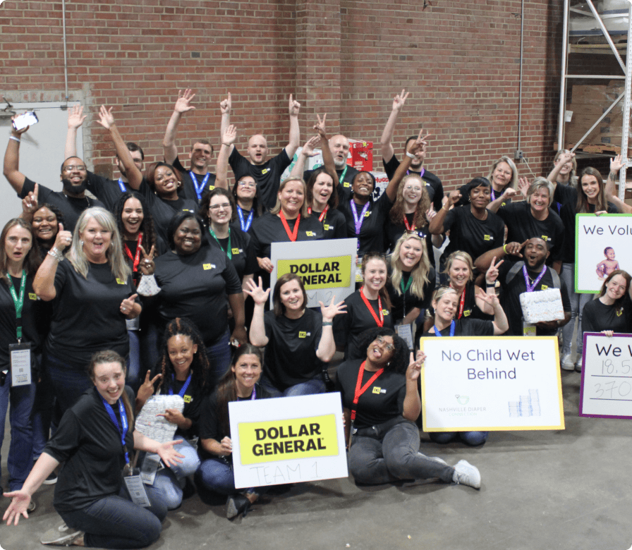 A large group of people in black shirts pose indoors, smiling and raising their hands. They hold signs reading “Dollar General,” “No Child Wet Behind,” and “We Volunteer.” The setting has brick walls in the background.