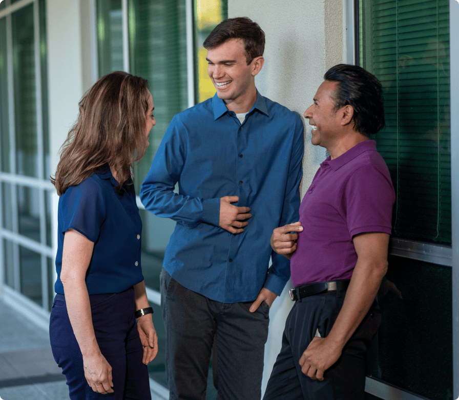 Three people stand and laugh together outside a building with large windows. They appear to be enjoying a light-hearted conversation. All are casually dressed and facing each other, smiling.