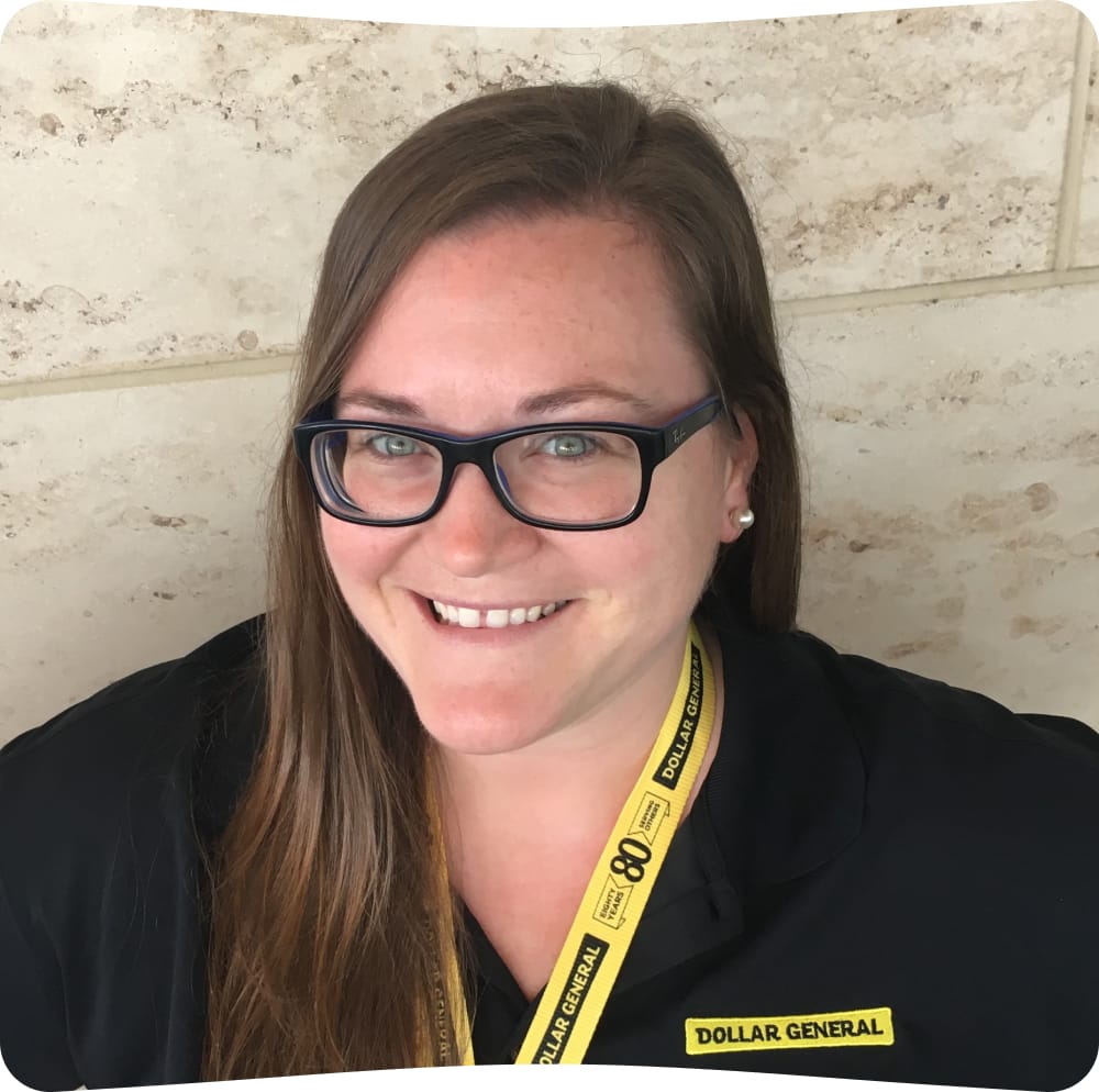 A woman with long brown hair and glasses smiles at the camera. She wears a black Dollar General shirt and a yellow lanyard. The background is a light-colored stone wall.