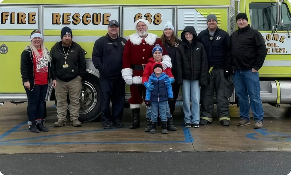 A group of people, including someone dressed as Santa Claus, pose in front of a yellow fire rescue truck. Two children stand in front, and the group is dressed warmly for cold weather.