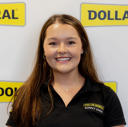 A woman with long brown hair, wearing a black “Dollar General Supply Chain” shirt, smiles in front of a white backdrop with multiple Dollar General logos.