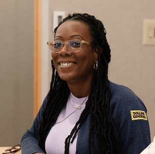A woman with long braids and glasses smiles while sitting at a table. She wears a blue Dollar General sweater over a lavender top and a necklace.
