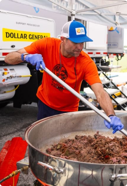 A man in an orange shirt and blue hat stirs a large pot of food outdoors under a tent, wearing blue gloves. A Dollar General sign and trailer are visible in the background.