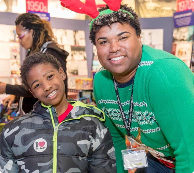 A smiling child in a camouflage jacket and a man in a festive green sweater with reindeer antlers pose together at a cheerful indoor event.
