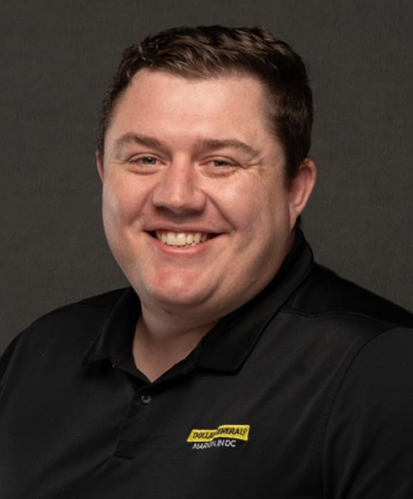 A smiling man with short brown hair wears a black collared shirt with a yellow and white “Duke Libraries” name tag, posed in front of a dark background.