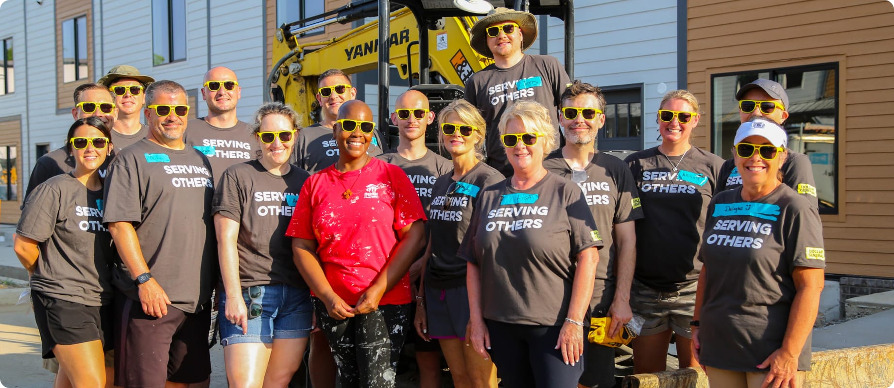 A group of volunteers wearing matching “Serving Others” shirts and yellow sunglasses pose and smile together outside at a construction site on a sunny day.