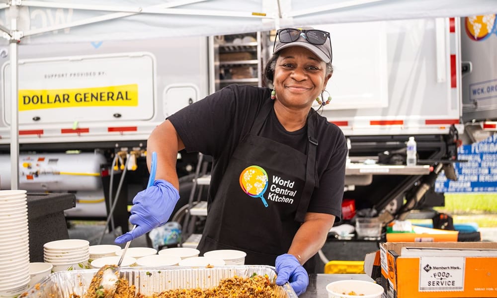 A smiling woman wearing gloves and a World Central Kitchen apron serves food from a large tray at an outdoor event, with bowls and food supplies on the table beside her.