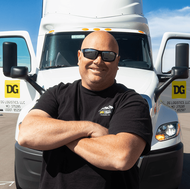 A smiling man wearing sunglasses and a black shirt stands with arms crossed in front of a white semi-truck with visible DG Logistics logos on the doors.