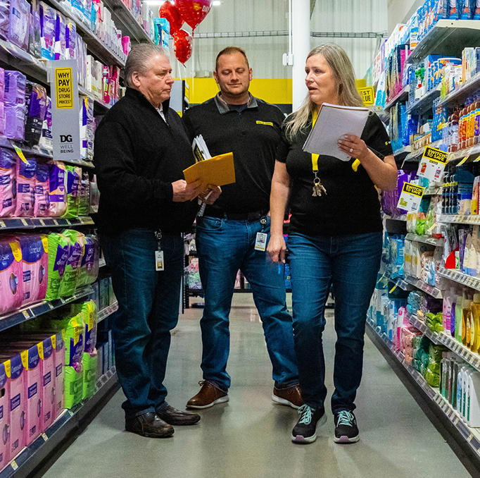 Three people wearing ID badges stand in a store aisle lined with products, holding clipboards and papers, appearing to discuss or inspect items on the shelves.