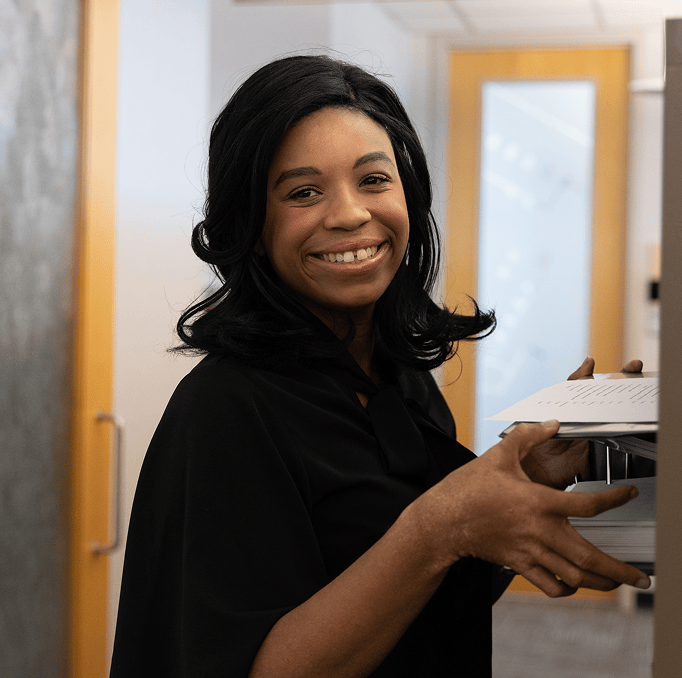 A woman with shoulder-length black hair smiles while holding a stack of papers or files in an office hallway with wooden doors and frosted glass.