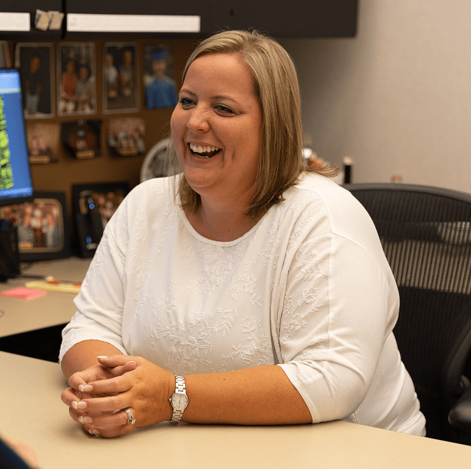 A woman in a white top sits at a desk, smiling and talking. Family photos and a computer monitor are visible in the background.