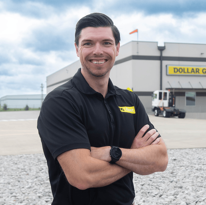 A man in a black polo shirt stands smiling with arms crossed outside a Dollar General building, with a truck and gravel in the background under a cloudy sky.