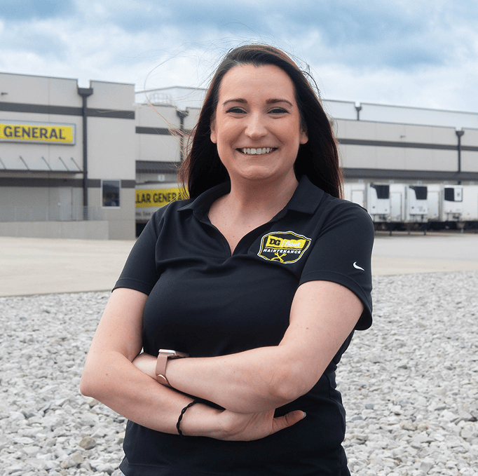 A woman in a black polo shirt stands smiling with arms crossed outside a Dollar General distribution center, with trucks and signage visible in the background.