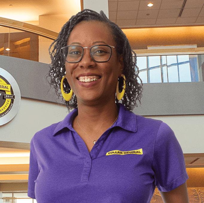 A smiling woman wearing glasses, yellow hoop earrings, and a purple Dollar General polo stands inside a brightly lit store with a modern interior and Dollar General signage in the background.