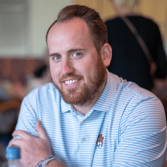 A man with a beard and short hair smiles at the camera. He is wearing a light blue and white striped polo shirt and is seated indoors with a blurred background.
