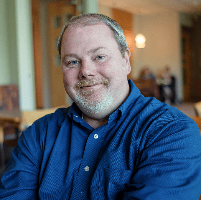 Smiling man with gray hair and beard wearing a blue shirt sits indoors with blurred people and furniture in the background.