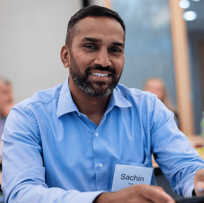 A man with a beard and mustache, wearing a blue shirt and a name tag reading “Sachin,” smiles while sitting at a table in a brightly lit room.
