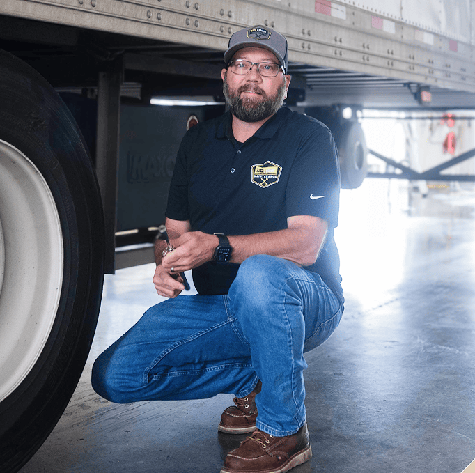 A man with a beard, glasses, and a cap kneels beside a semi-truck trailer indoors. He wears a black polo shirt, blue jeans, and brown boots, looking at the camera and smiling slightly.