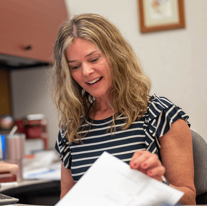 A woman with wavy blonde hair wearing a striped shirt sits at a desk, smiling as she looks at documents in her hand.
