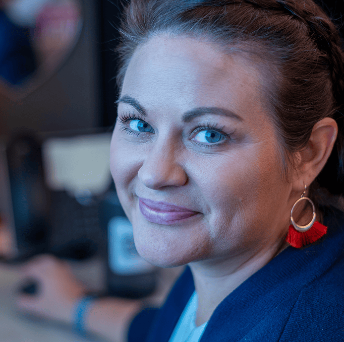 A woman with blue eyes, light skin, and brown hair smiles at the camera. She wears red tassel earrings and a blue top, sitting at a desk with a blurred background.