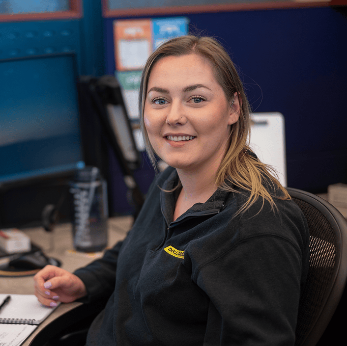 A woman with blonde hair sits at a desk in an office, smiling at the camera. She wears a black fleece jacket and holds a pen above a notebook. Computer monitors and office supplies are visible in the background.