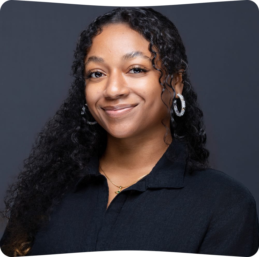 Woman with long curly hair, wearing hoop earrings and a black collared shirt, smiles gently against a dark background.