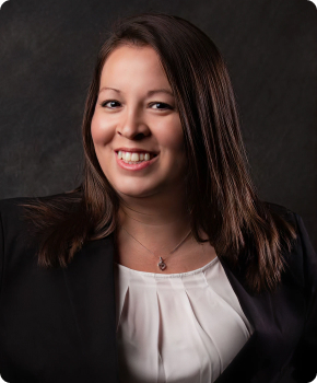 A woman with long brown hair, wearing a black blazer over a white blouse, smiles at the camera against a dark background.