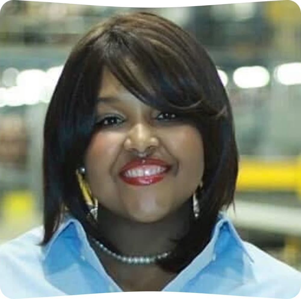 A woman with straight dark hair, wearing a light blue collared shirt and a pearl necklace, smiles at the camera with a blurred industrial background behind her.