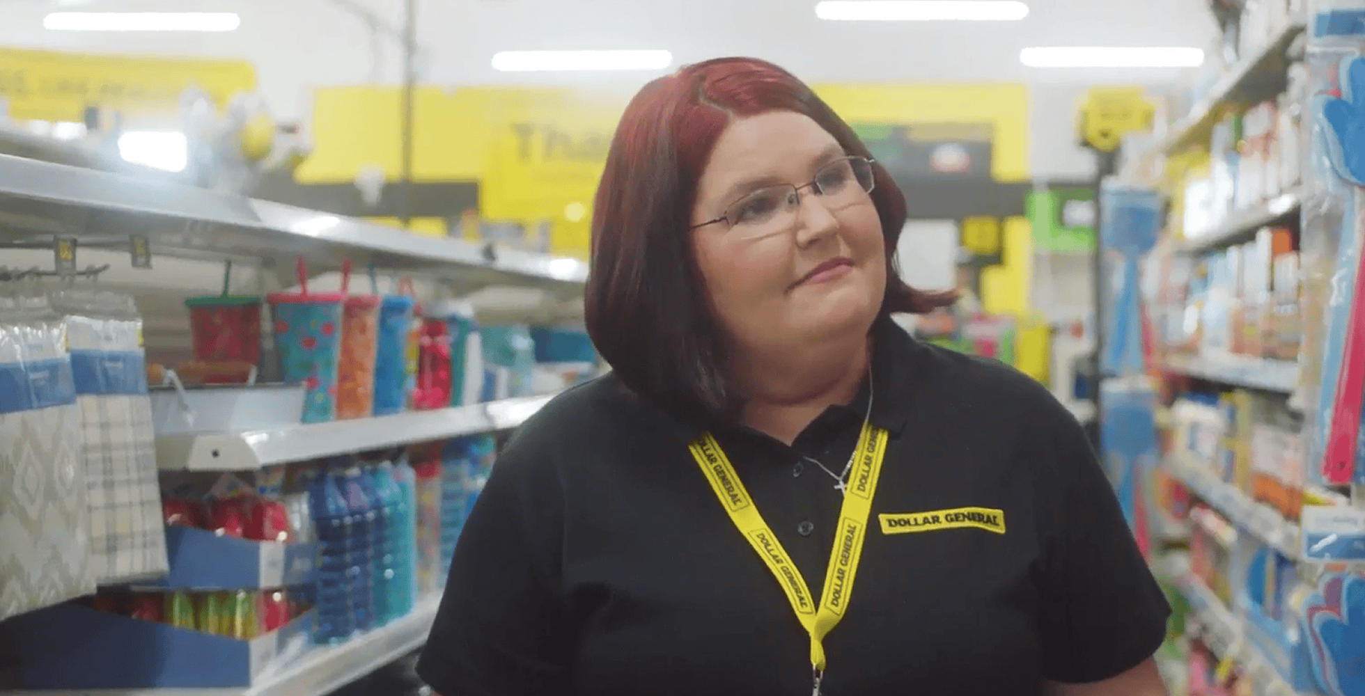 A woman with red and brown hair, wearing glasses and a Dollar General uniform, stands in an aisle of a brightly lit store with party supplies on the shelves.