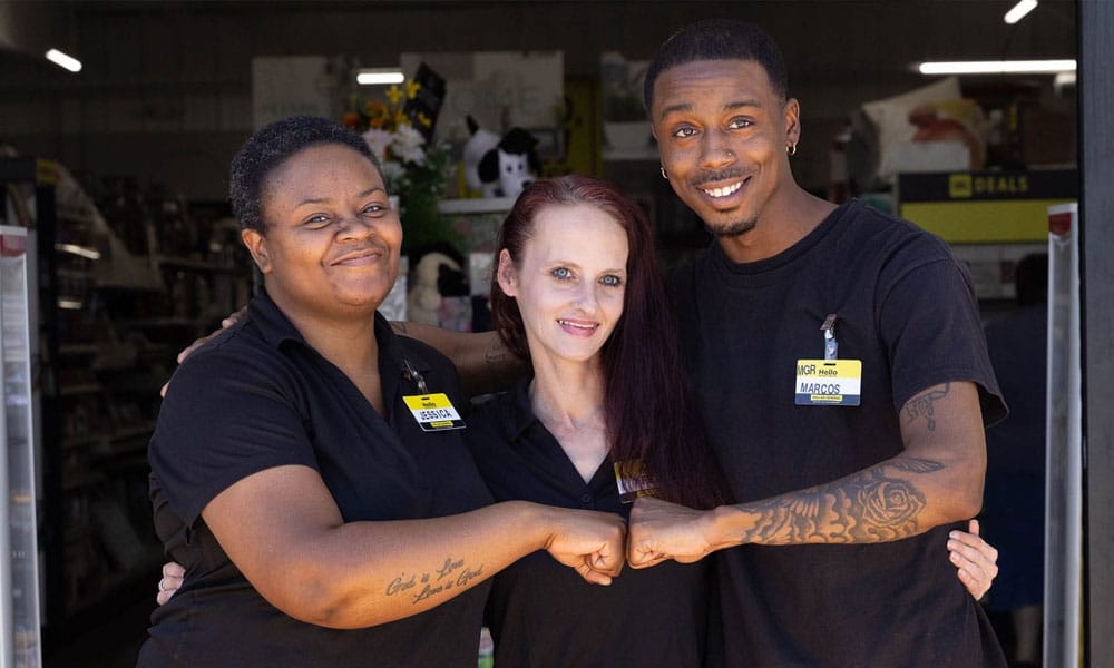 Three smiling store employees in black shirts stand together in front of a store, arms around each other, with two of them fist-bumping. All wear nametags and appear happy and united.