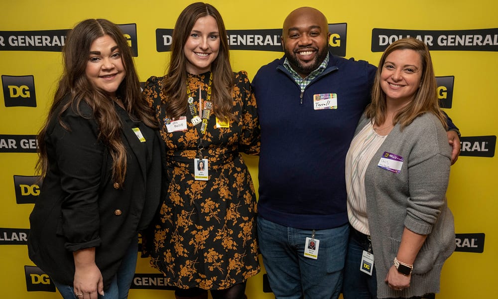 Four smiling people stand in front of a yellow Dollar General backdrop, wearing name tags and business-casual clothes, posing together with arms around each other.