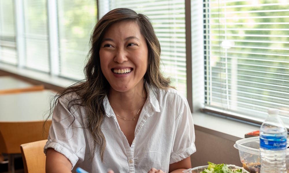 A woman in a white shirt smiles while sitting at a table with food, including a salad and a water bottle, near large windows with sunlight streaming in.