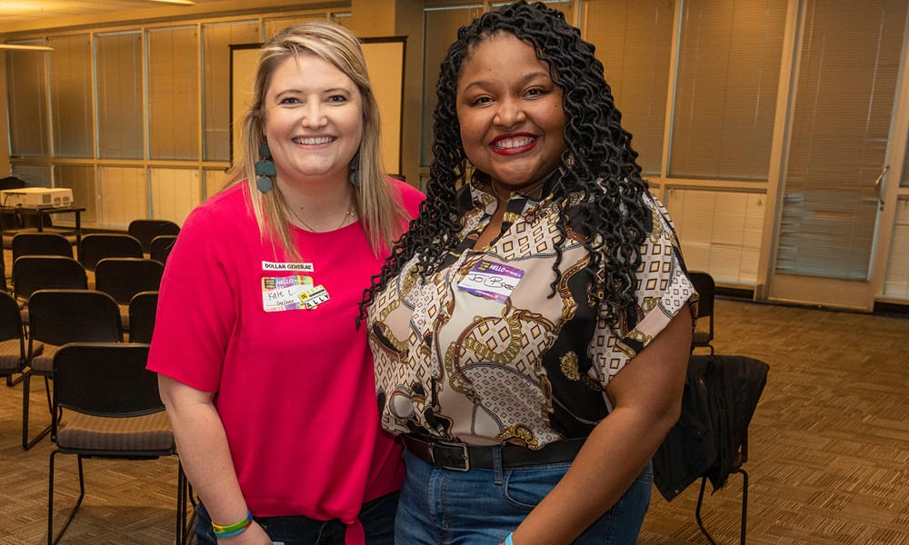 Two women smiling and standing close together in a room with empty chairs. Both are wearing nametags and casual outfits, with one in a bright pink shirt and the other in a patterned blouse.