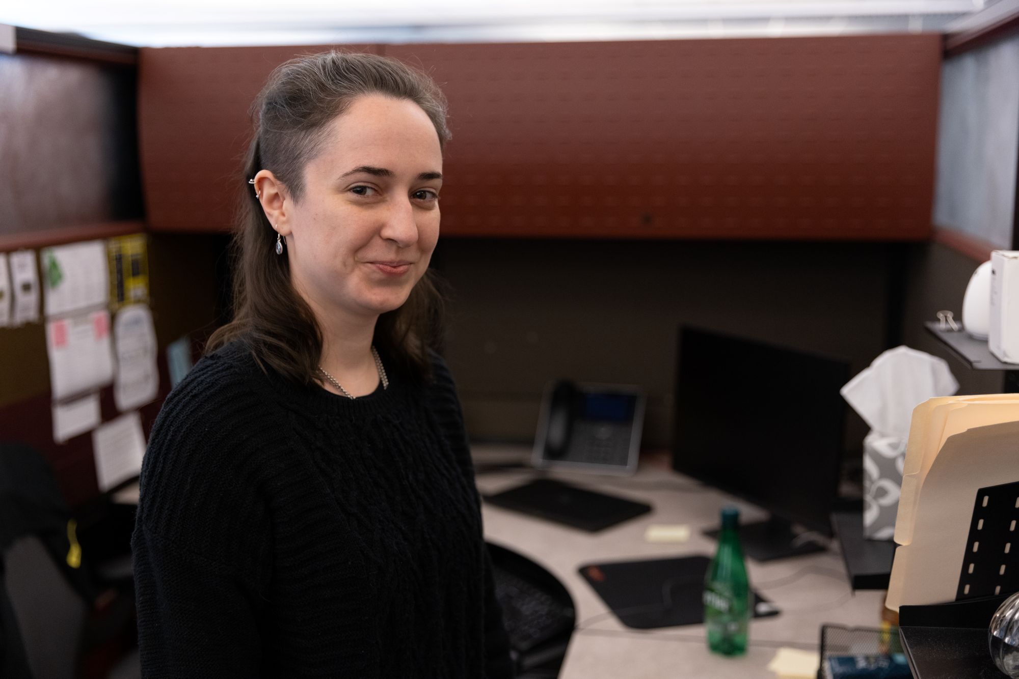A woman wearing a black sweater smiles while standing in a cubicle office.